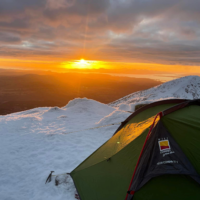 A green backpacking tent staked into snow on a mountain ridge at sunrise, warm orange light spilling across the snowy landscape and distant islands under a cloudy sky with an ice axe planted nearby.