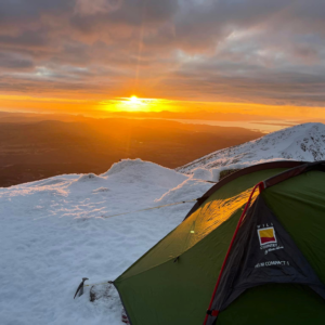 A green backpacking tent staked into snow on a mountain ridge at sunrise, warm orange light spilling across the snowy landscape and distant islands under a cloudy sky with an ice axe planted nearby.
