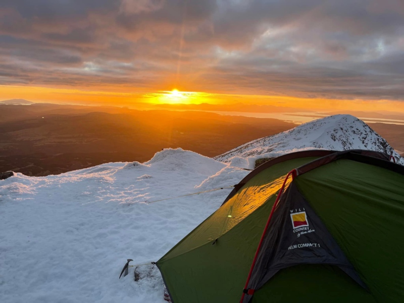 ls_wc_sunset_mountian_helm-Compact-1_2022 A green backpacking tent staked into snow on a mountain ridge at sunrise, warm orange light spilling across the snowy landscape and distant islands under a cloudy sky with an ice axe planted nearby.