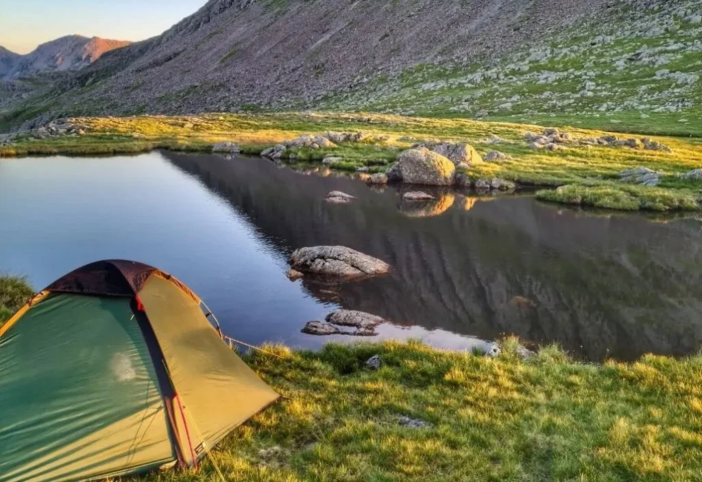 Small green tent pitched on a grassy bank beside a calm mountain tarn that reflects rocky slopes and scattered boulders in warm golden light