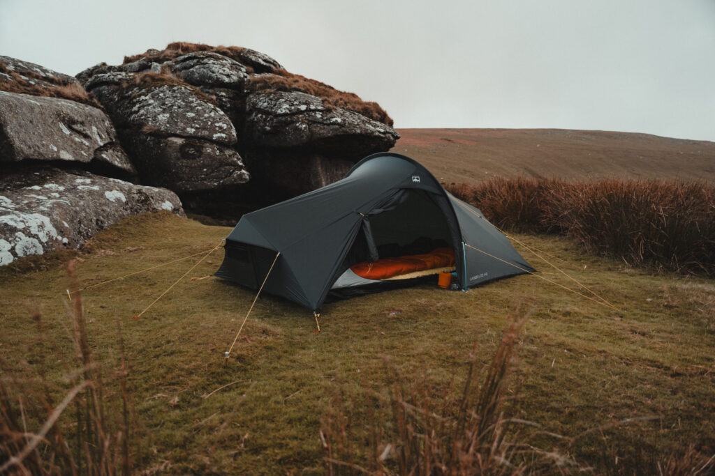 Lightweight yellow one-person tent pitched on a windswept grassy hillside with rocky mountains and an overcast sky in the background