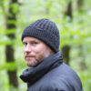 134-square-1.jpg Bearded man wearing a dark grey knitted beanie with a small logo tag and a black puffer jacket, looking over his shoulder against a blurred green woodland background.