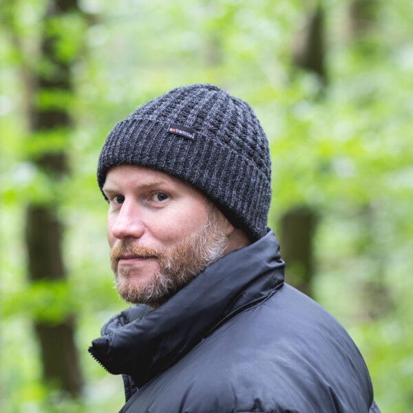 134-square-1.jpg Bearded man wearing a dark grey knitted beanie with a small logo tag and a black puffer jacket, looking over his shoulder against a blurred green woodland background.