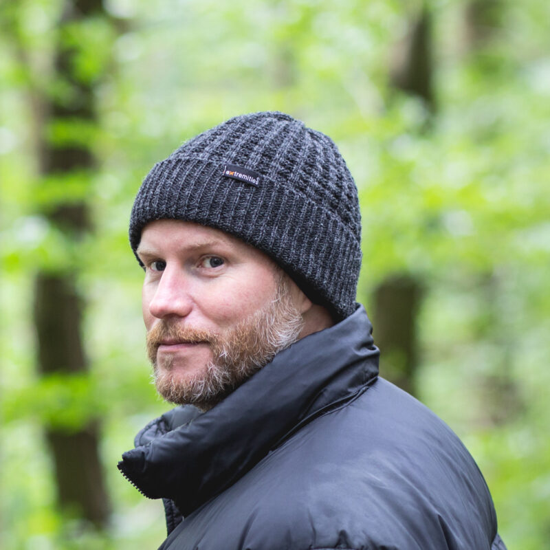 134-square-1.jpg Bearded man wearing a dark grey knitted beanie with a small logo tag and a black puffer jacket, looking over his shoulder against a blurred green woodland background.