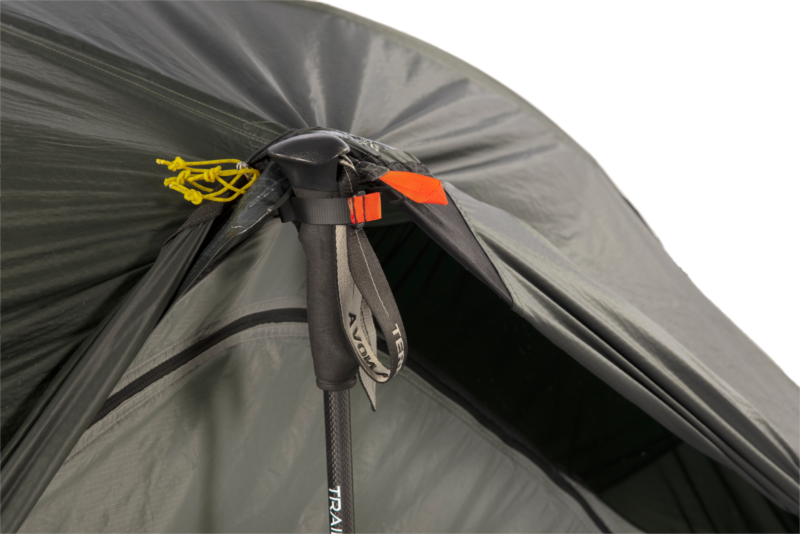 Close-up of a grey-green tent apex where a black trekking pole with foam grip and wrist strap props open the fly, secured by an orange fabric tab and knotted yellow guy lines.