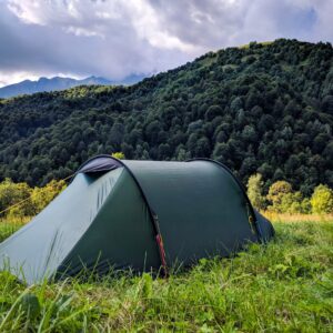 Dark green tunnel tent pitched on a grassy meadow in the foreground with dense tree-covered hills and distant mountain peaks beneath a dramatic cloudy sky.