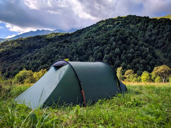 Dark green tunnel tent pitched on a grassy meadow in the foreground with dense tree-covered hills and distant mountain peaks beneath a dramatic cloudy sky.