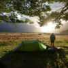 Green solo tent pitched on a grassy riverside plain at sunrise, a camper standing beside scattered gear under overhanging tree branches with distant mountains silhouetted against the bright low sun.