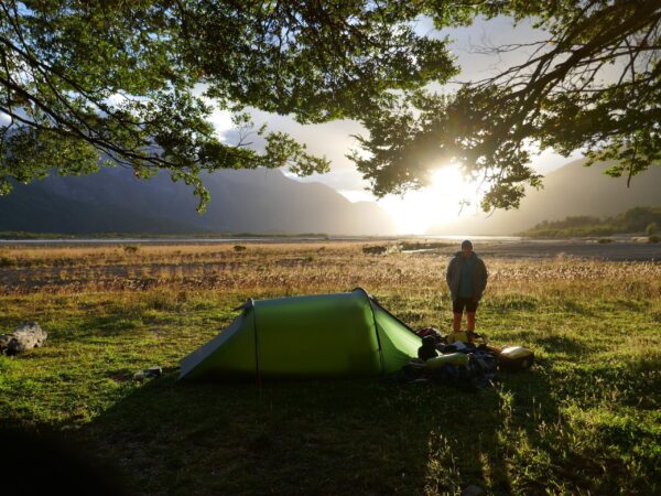 Green solo tent pitched on a grassy riverside plain at sunrise, a camper standing beside scattered gear under overhanging tree branches with distant mountains silhouetted against the bright low sun.