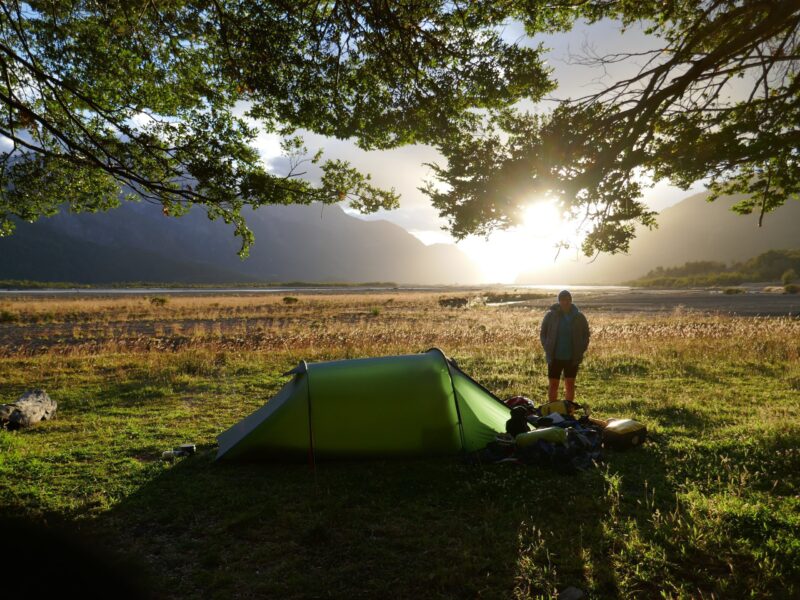 2019sa45-1-scaled-2.jpg Green solo tent pitched on a grassy riverside plain at sunrise, a camper standing beside scattered gear under overhanging tree branches with distant mountains silhouetted against the bright low sun.