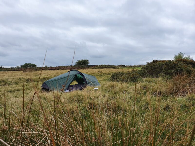 20200910_124151-scaled-2.jpg A small green camping tent pitched among tall windswept grasses and low scrub on an overcast grassy moor beneath a cloudy grey sky.