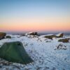 20221215_154614-scaled-2.jpg A small green tent pitched on snow-dusted grass amid large frost-covered granite boulders on a windswept moor beneath a pastel pink and blue sunrise sky.