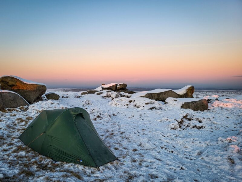 20221215_154614-scaled-2.jpg A small green tent pitched on snow-dusted grass amid large frost-covered granite boulders on a windswept moor beneath a pastel pink and blue sunrise sky.