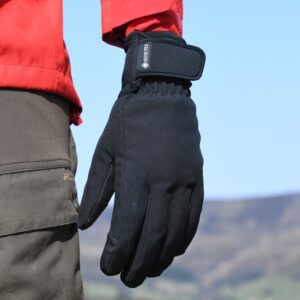 Close-up of a black GORE-TEX winter glove with a Velcro wrist strap worn beside a red jacket sleeve and brown trousers, set against a clear blue sky and distant hills.