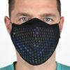 23MRFM-Maze-Runner-Face-Mask-Worn-Daylight-1500x1500px-1.jpg Close-up of a man wearing a black cloth face mask with an iridescent hexagonal honeycomb pattern, showing his light blue eyes and short dark hair against a plain background.