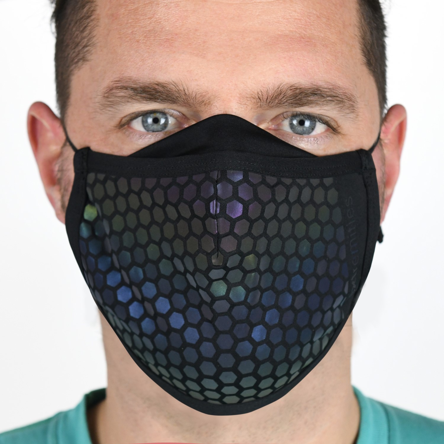 23MRFM-Maze-Runner-Face-Mask-Worn-Daylight-1500x1500px-1.jpg Close-up of a man wearing a black cloth face mask with an iridescent hexagonal honeycomb pattern, showing his light blue eyes and short dark hair against a plain background.