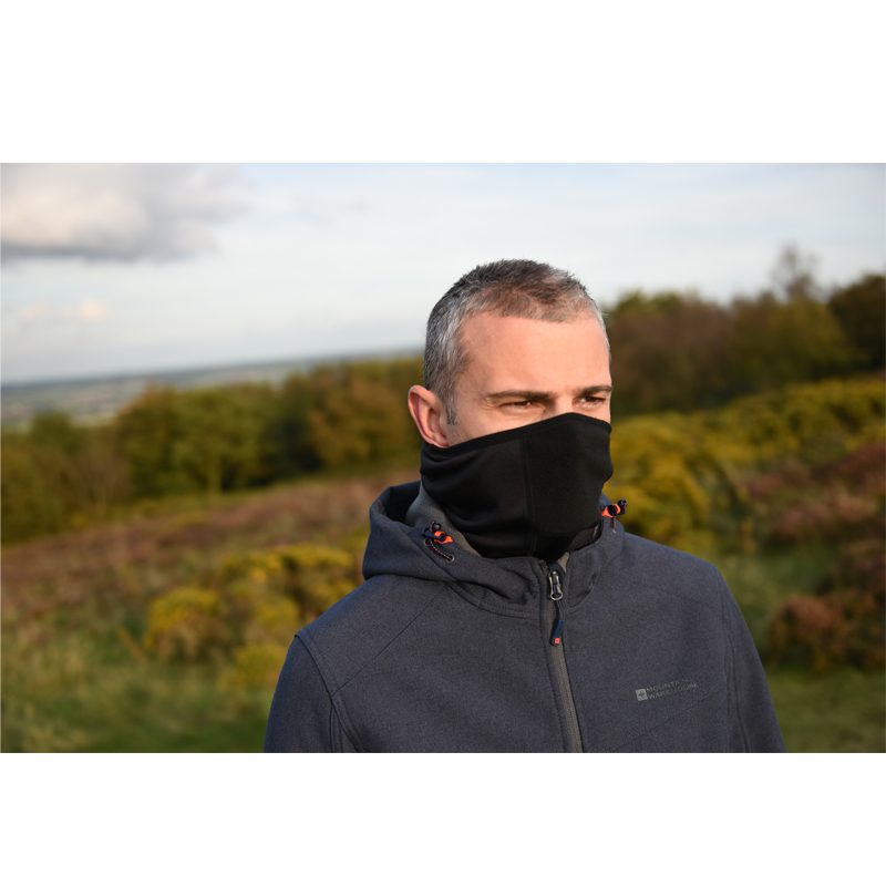 Man with short grey hair wearing a black face gaiter and a dark hooded jacket, standing on a grassy hillside with shrubs and trees under a cloudy sky