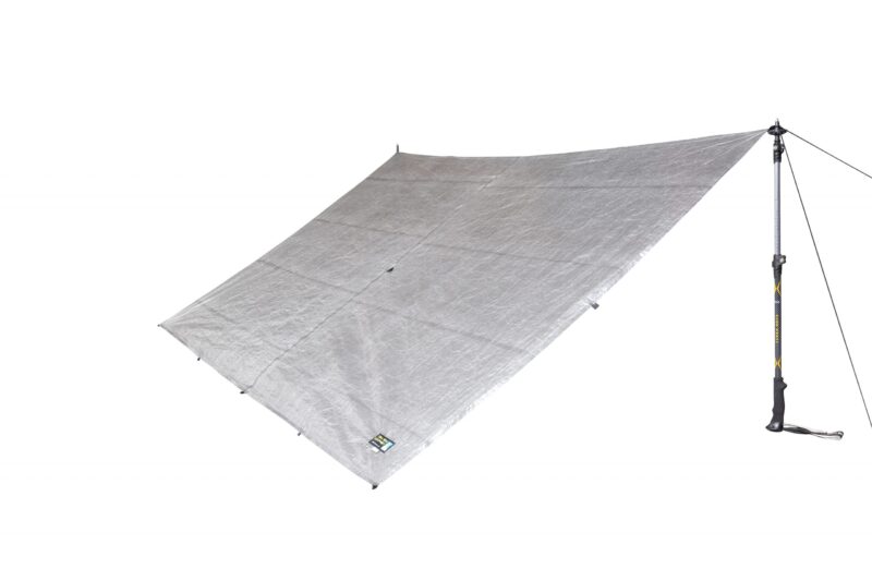 43TU1-Tarp-Ultra-1-Back-View-scaled-4.jpg Rectangular silver camping tarp pitched at an angle and anchored by a trekking pole and guyline, showing reinforced corner tabs and a small brand label on a plain white background.