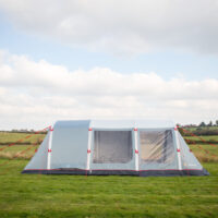 Large light-grey family tent with red guy ropes and clear plastic windows pitched on a grassy field with rolling hills and a cloudy sky in the background.