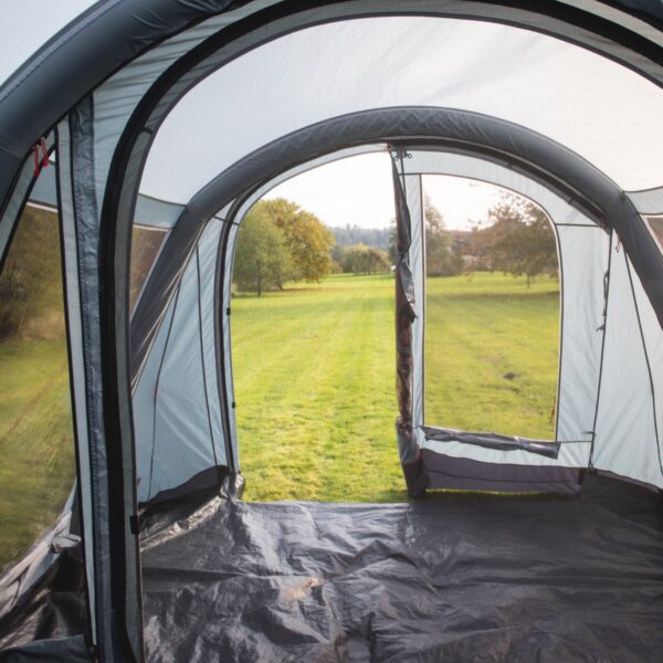 44ZON4E-Zonda-4EP-view-out-of-front-1500x1500-1-1.jpg Inside a grey inflatable tent with a black groundsheet, the open doorway frames a mown green field with scattered trees and a distant treeline under a pale sky.