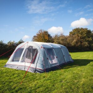 Large grey tunnel-style family tent with red guy ropes and mesh front windows pitched on a green grassy field with a hedgerow and trees beneath a blue sky dotted with white clouds