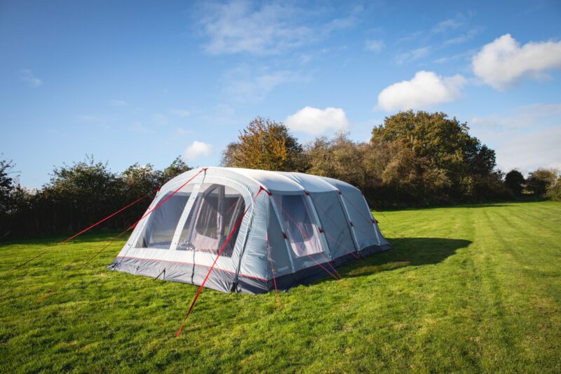 Large grey tunnel-style family tent with red guy ropes and mesh front windows pitched on a green grassy field with a hedgerow and trees beneath a blue sky dotted with white clouds