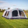 A large grey and navy family tent with arched roof, red guy lines and multiple mesh windows pitched on a sunlit green field with rolling countryside and trees in the background.