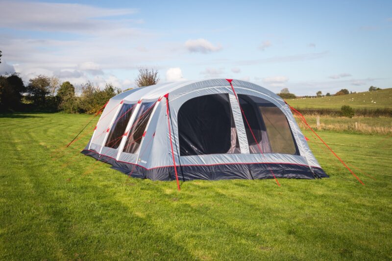 A large grey and navy family tent with arched roof, red guy lines and multiple mesh windows pitched on a sunlit green field with rolling countryside and trees in the background.