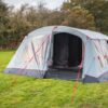 Large light-grey tunnel-style family tent with red guy ropes staked into a grassy field, open front revealing a dark interior and clear window panels, with trees and bushes in the background.