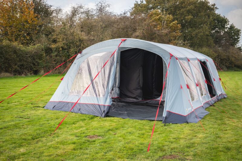 Large light-grey tunnel-style family tent with red guy ropes staked into a grassy field, open front revealing a dark interior and clear window panels, with trees and bushes in the background.