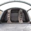 Spacious grey inflatable tent interior with arched air beams, a central dark sleeping pod with two partially open zip doors and red-trimmed hanging storage pockets, and a tarp-covered floor opening onto a grassy field.