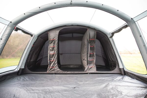 Spacious grey inflatable tent interior with arched air beams, a central dark sleeping pod with two partially open zip doors and red-trimmed hanging storage pockets, and a tarp-covered floor opening onto a grassy field.