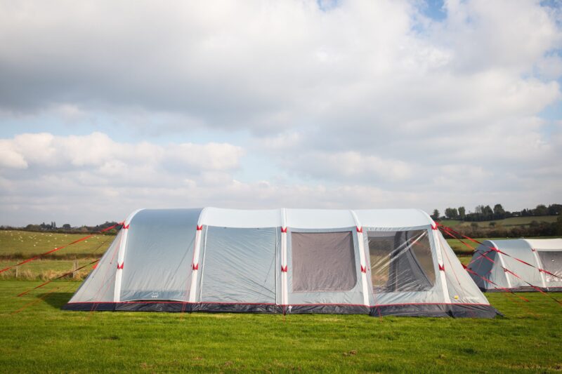 Large grey and white tunnel-style family tent with red guy ropes pitched on a green field under a cloudy sky, with rolling hills and another tent visible in the background.