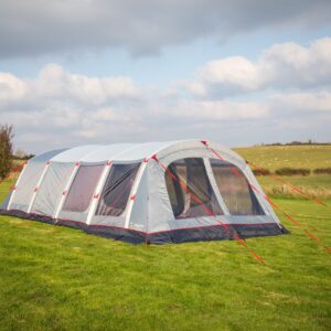 Large grey and navy tunnel-style family tent with mesh windows and bright red guy lines staked on a green grassy field under a partly cloudy sky, with rolling farmland and hedges in the background.