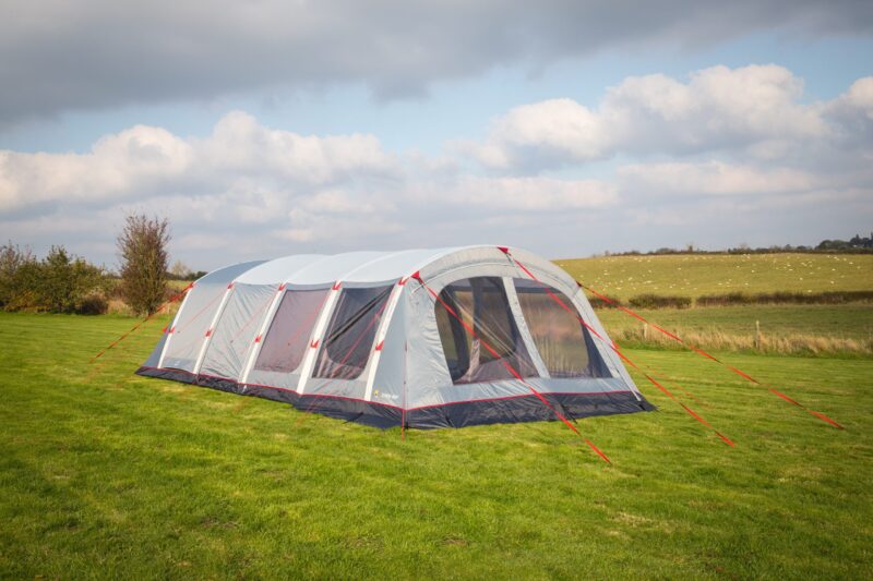 Large grey and navy tunnel-style family tent with mesh windows and bright red guy lines staked on a green grassy field under a partly cloudy sky, with rolling farmland and hedges in the background.