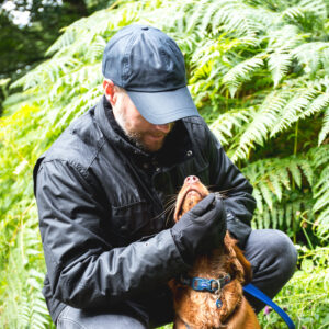 Bearded man in a black cap and jacket crouching amid green ferns, gently holding the chin of a brown dog with a blue collar as the dog tilts its head up.