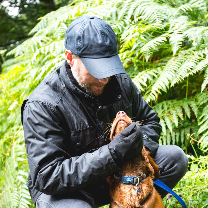 Bearded man in a black cap and jacket crouching amid green ferns, gently holding the chin of a brown dog with a blue collar as the dog tilts its head up.
