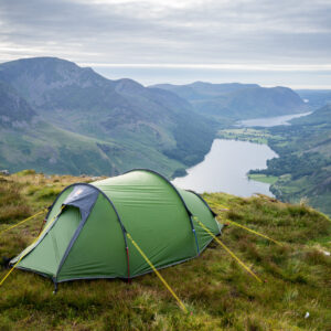 Green tunnel tent pegged with yellow guy lines on a grassy hilltop, overlooking a long lake winding through steep, green mountains under a cloudy sky