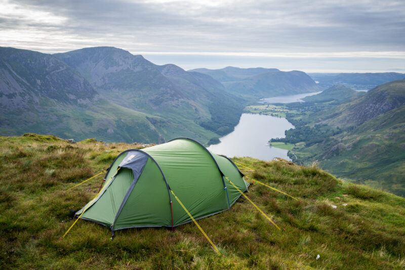 Green tunnel tent pegged with yellow guy lines on a grassy hilltop, overlooking a long lake winding through steep, green mountains under a cloudy sky