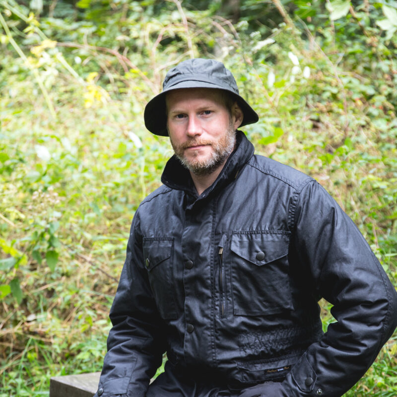 63-square-1.jpg Bearded man wearing a black bucket hat and dark jacket sitting outdoors on a wooden bench amid dense green foliage, looking toward the camera.
