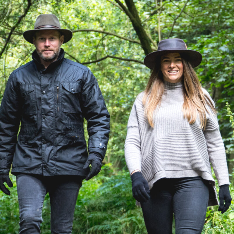 69-square-1.jpg A man and woman wearing wide-brimmed hats, gloves and outdoor clothing walk side by side along a green wooded path, the woman smiling in a grey ribbed sweater and the man in a black jacket.