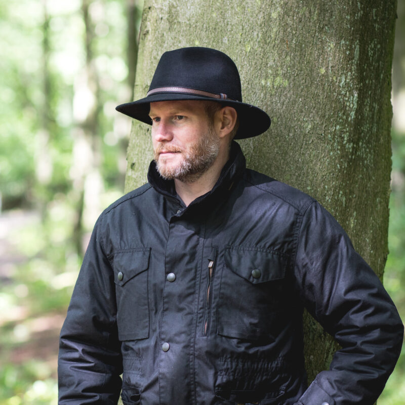 Bearded man wearing a black wide-brimmed hat and dark jacket leaning against a tree in a sun-dappled forest, looking thoughtfully to the left.