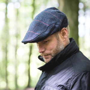 Profile of a bearded man looking down, wearing a blue checked tweed flat cap and a black jacket against a softly blurred woodland background