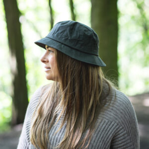 Woman in profile wearing a dark bucket hat and a grey ribbed sweater, standing among sunlit trees with long brown hair falling over her shoulders