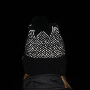 Rear view of a person wearing a black knitted pompom hat with a white reflective zigzag pattern and a turned-up ribbed brim, black jacket collar and a few strands of hair visible against a dark background.