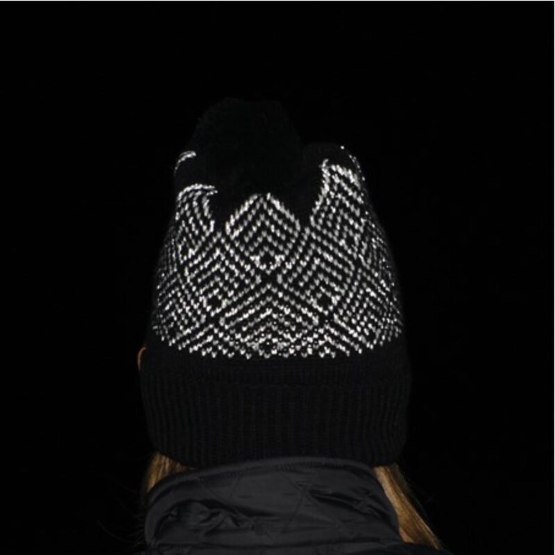 Antares-Beanie-Dark-1500px-96dpi-1.jpg Rear view of a person wearing a black knitted pompom hat with a white reflective zigzag pattern and a turned-up ribbed brim, black jacket collar and a few strands of hair visible against a dark background.