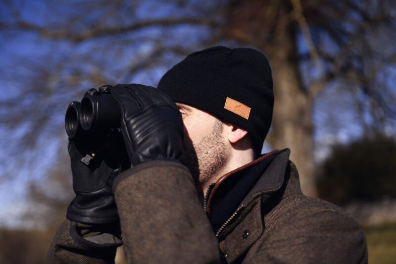 Man in a black beanie and leather gloves holding binoculars to his eyes while wearing a brown winter coat outdoors against a blurred background of bare trees.