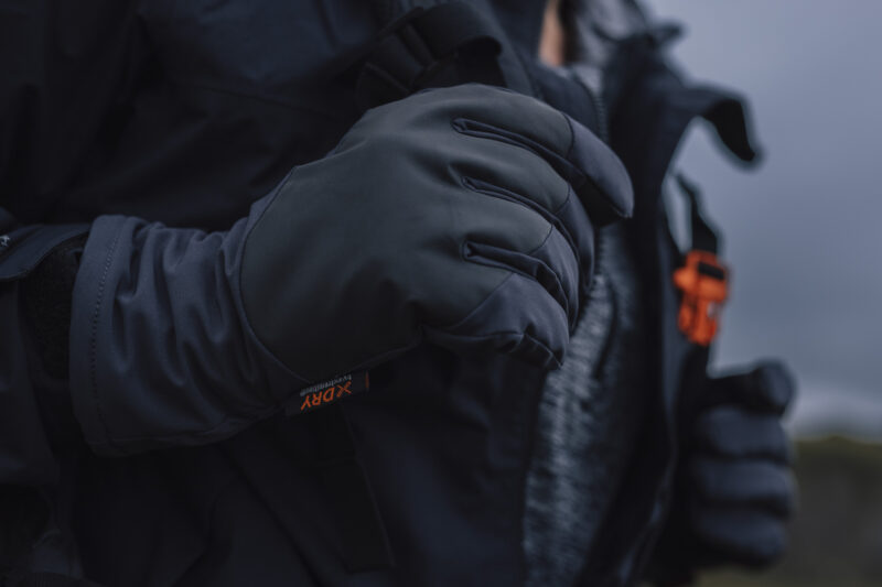 Aurora Gloves AW25 (4) Close-up of a person's hand wearing a black outdoor glove, showcasing its stylish design and functionality for cold weather activities. The glove is part of a weather-resistant jacket ensemble, ideal for hiking or winter sports.