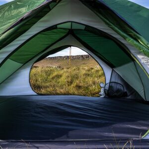 Interior view of a green-and-white dome tent with the entrance unzipped, the opening framing a grassy meadow and low rocky ridge under a blue sky.