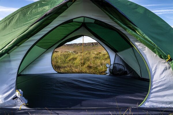 Interior view of a green-and-white dome tent with the entrance unzipped, the opening framing a grassy meadow and low rocky ridge under a blue sky.